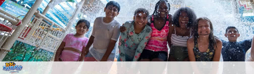 Image shows a group of happy children in swimwear standing under a water splash structure at Big Kahuna's Water Park. They're smiling and enjoying the water cascading around them. The park's logo is visible in the corner.