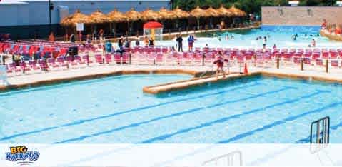 A vibrant outdoor swimming pool scene with clear blue water. Multiple swimmers enjoy the facility alongside rows of red lounge chairs under a sunny sky. A hut with a thatched roof provides a shaded area in the distance, adding a tropical vibe to the ambience. Safety is ensured with lifeguards and poolside ladders.