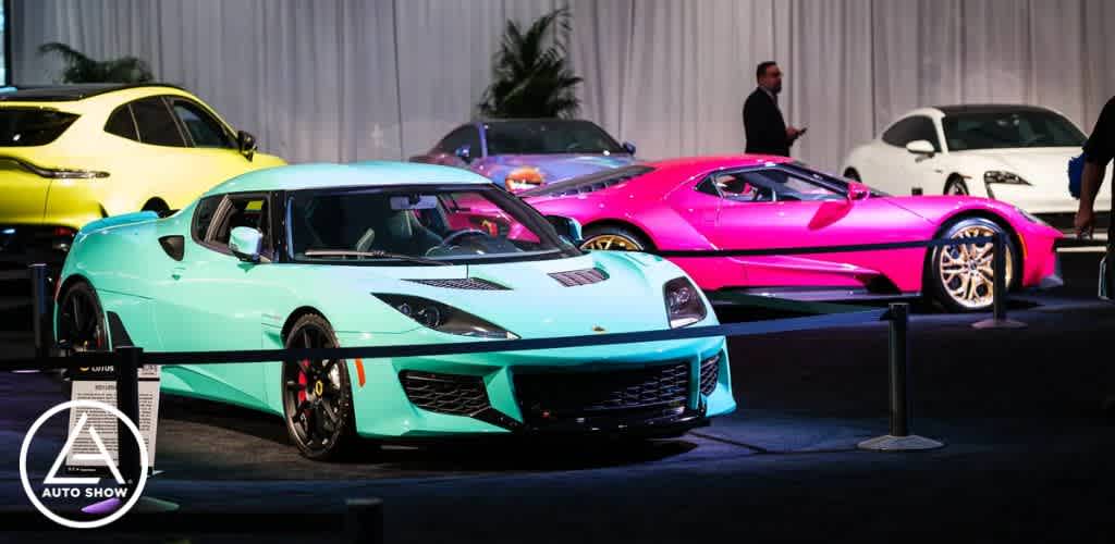 This image displays a vibrant scene from an auto show where three luxury sports cars are positioned prominently in the foreground under bright exhibition lighting. From left to right: a glossy yellow car partially visible, a turquoise sports car with a black roof and accents, and a hot pink sports car with a shiny gold rimmed wheel. These sleek vehicles are on display behind a stanchion, set against a muted backdrop of other vehicles and event attendees. One individual is observed standing by the pink car, seemingly engaged in conversation. In the background, other people are visible, walking or standing near the cars in a spacious exhibition hall adorned with drapery. A logo for the auto show is present in the lower left corner of the image.

Find your next great adventure at GreatWorkPerks.com, where we offer the convenience of getting your tickets at the lowest prices, ensuring your savings put you in the fast lane to fun!