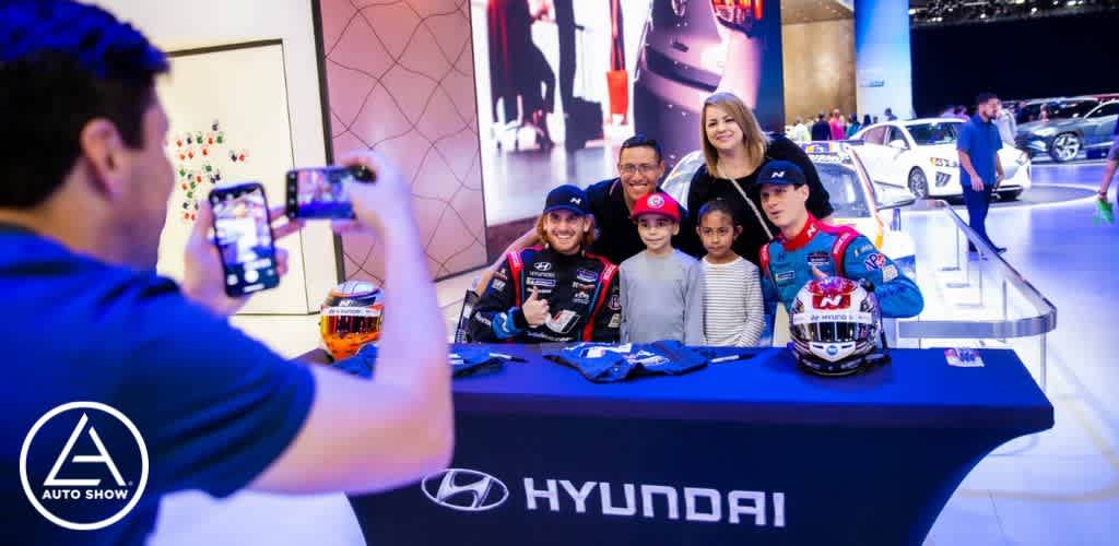 Image description:

This is a vibrant photograph taken at an auto show, featuring a Hyundai exhibition booth. In the foreground, on the left side, an individual is capturing the moment with a smartphone, taking a photo of a group standing behind a table adorned with the Hyundai logo. The group includes two individuals dressed in racing attire complete with sponsorship patches and sitting on bar stools on the left side of the table. They are joined by two adults standing and two children, all smiling towards the camera. Everyone appears joyful as they engage in this photo opportunity. The background shows several cars on display with soft focus and bright exhibition lighting, creating a lively ambiance typical of such events. The logo of the Auto Show is visible in the bottom left corner of the image, representing the event where this moment is taking place.

Experience the thrill of the Auto Show and capture memories without breaking the bank; visit GreatWorkPerks.com for the lowest prices and significant savings on tickets.