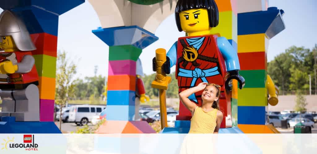 Image shows a smiling child in a yellow top posing playfully with a large LEGO figurine dressed as a knight outside the colorful LEGOLAND Hotel. Bright blue skies and a parking area in the background complement the cheerful setting.