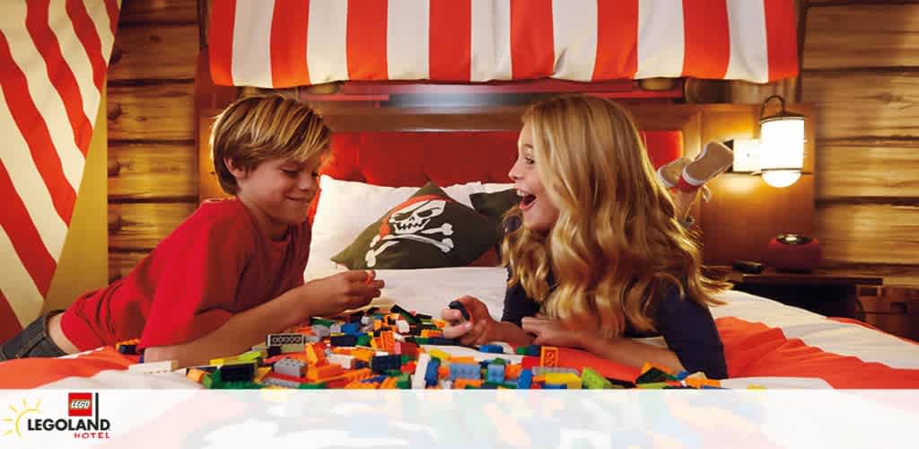 Two children are playing with colorful LEGO bricks on a bed in a wood-paneled room with a pirate-themed decor. The bed has striped red and white bedding, and there's an atmosphere of joy and playfulness. The LEGOLAND Hotel logo is visible in the bottom left corner, suggesting this is part of a themed hotel experience.