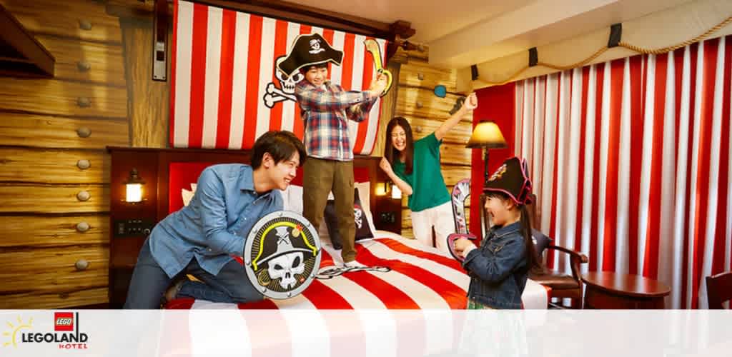 A family enjoys a pirate-themed room at LEGOLAND Hotel. A child wearing a pirate hat stands on a bed with red and white stripes, holding a sword. Another child kneels with a shield. An adult cheers on, and another grins while watching the playful scene. Cabin-like walls add to the adventurous ambiance.