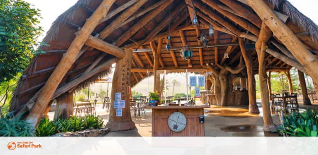 Description: The image features an expansive outdoor pavilion with a rustic design, located at the San Diego Zoo Safari Park. The structure is built with large wooden beams and pillars that support a multi-tiered thatched roof, offering a shaded area for visitors to relax. Beneath the roof, there are several round tables with chairs arranged for dining or resting. To the left, there's a significant wooden pillar with engravings that appears to be a structural component as well as a decorative element. A variety of hanging lanterns add to the ambiance. The flooring consists of a smooth concrete surface, and lush green plants edge the pavilion, blending the man-made structure with the natural surroundings. There is a clock visible at the bottom of the image, indicating the consideration for guests to keep track of time during their visit. Experience the thrills of the wild and make sure to take advantage of our exclusive discounts on tickets for a memorable adventure at the lowest prices available!