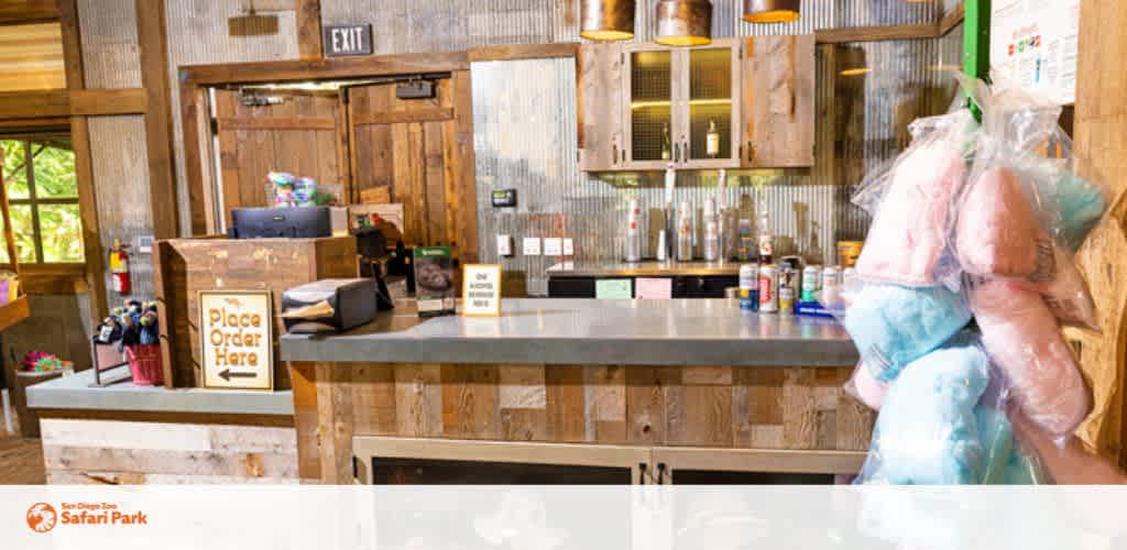 Description: This image depicts the interior of a casual service area with a rustic aesthetic at the San Diego Zoo Safari Park. The space features a wooden counter where the signage indicates a place to order with the words "Place Order Here." On the left side of the counter, there are umbrellas stored in a holder and a point-of-sale system for transactions. The back wall is lined with various vertical wooden planks, creating a warm and textured look, and in the background, metal industrial-style pendant lights hang from the ceiling. To the right, there are shelves stocked with condiment bottles and a glass display case near the exit sign. In the foreground, a bulk of what appears to be packaged cotton candy in pink and blue is partially visible. Please note that at FunEx.com, we strive to offer our customers exclusive discounts, savings with the lowest prices on tickets, ensuring an unforgettable experience at attractions like the Safari Park.