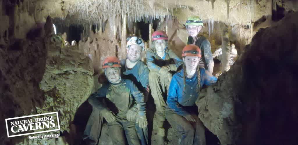 Group of six people in caving gear with helmets and lights pose inside a cavern. The environment is dark with limestone formations in the background. Everyone is smiling, appearing muddy from the cave exploration. The Natural Bridge Caverns logo with the text  Naturally Amazing  is visible in the corner.