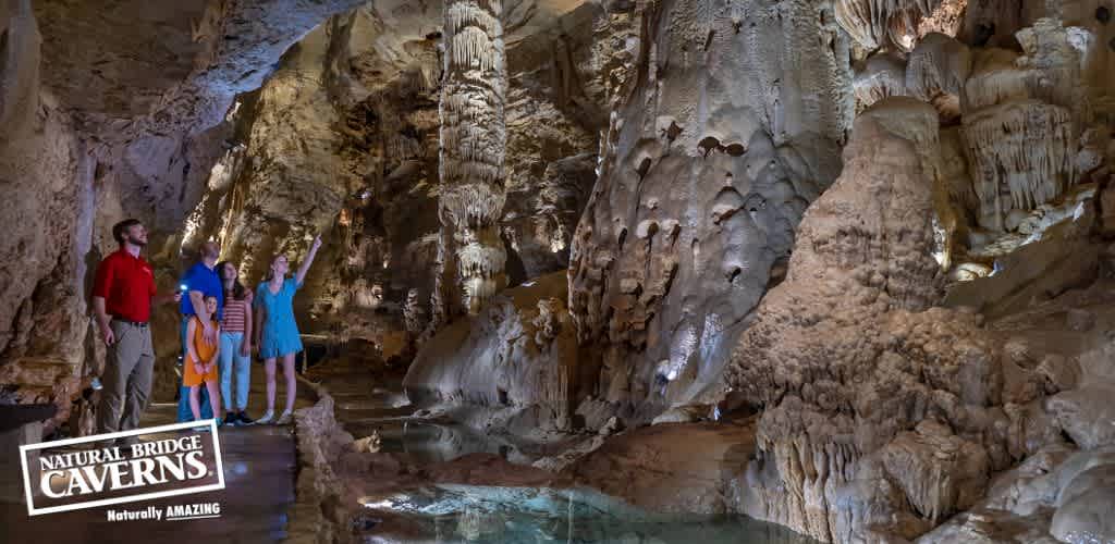 Image shows a group of people, including children, inside the naturally lit Natural Bridge Caverns. They stand on a path next to a tranquil water pool, looking up in wonder at the towering, intricately formed stalactites and stalagmites surrounding them. The cavern's sign is visible, indicating its name.