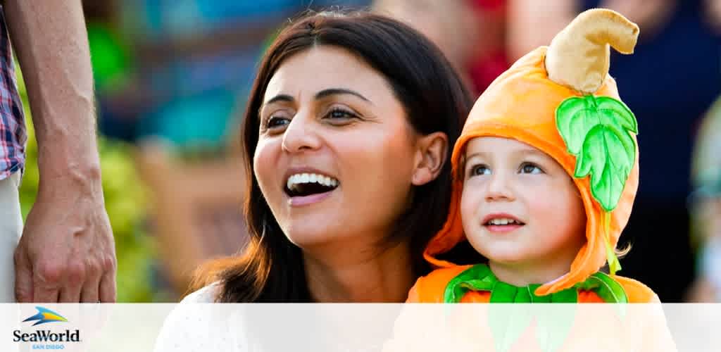 A cheerful woman and a child wearing an orange pumpkin costume with a hat shaped like a pumpkin stem look happily off to the side. The SeaWorld San Diego logo is visible in the lower left corner, suggesting the setting is a theme park. The focus is on their joyous expressions, capturing a moment of delight.