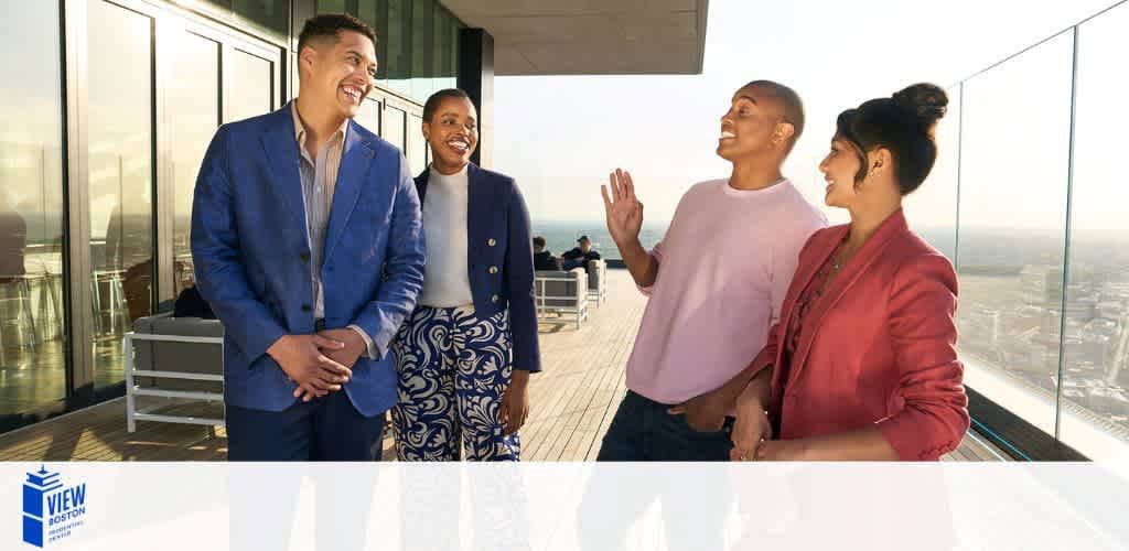 Image of four individuals engaging in a cheerful conversation on a high-rise building's outdoor terrace. They are dressed in smart casual and business attire, with a clear sky behind them, suggesting a pleasant day. The terrace overlooks a cityscape, indicated by the blurred buildings in the background. The logo on the bottom left corner reads 'VIEW Boston'.