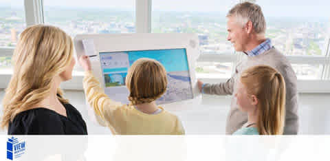 Image shows three individuals, possibly a family, standing in front of a window with a cityscape view. A man on the right holds up a landscape-oriented poster showing a beach scene, pointing something out to a young girl next to him. Another young girl and a woman on the left watch attentively. The room appears bright and modern.