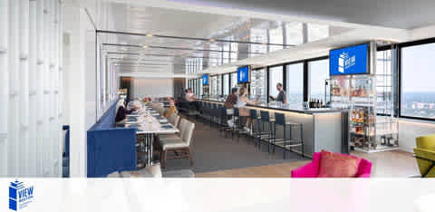 A modern office break room with a long bar and stools on the right, facing large panoramic windows that offer a city view. People are seated, working and socializing. Logo of The View is displayed on the wall and a fridge. Bright, airy ambiance.