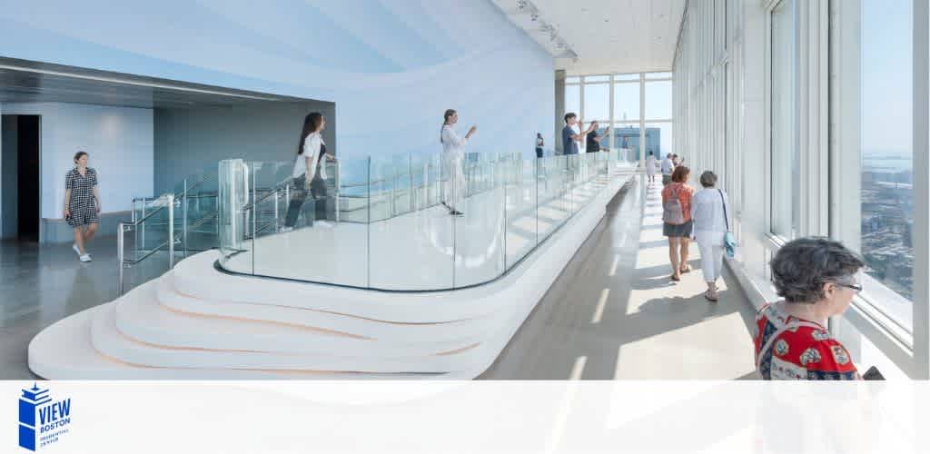 This image shows visitors at an indoor observation deck with clear glass barriers. People are walking around or standing, enjoying the view. The space is bright and airy with a high ceiling, and sunlight filters through large windows offering a panoramic cityscape. There's a logo in the bottom left corner reading 'VIEW Boston.'