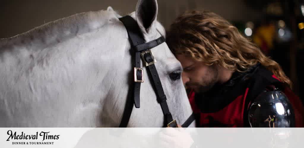 This image depicts a tender moment between a man and a horse, evoking the spirit of medieval camaraderie. The man, who appears to be a knight dressed in a red and black outfit with long curled hair, gently rests his forehead against the horse's, showing a strong bond between the knight and his trusted steed. The horse, with a glistening white coat and a black bridle, stands calmly during this moment of connection. Both are partially visible while the background fades into a soft blur, highlighting the intimacy of the interaction. The logo for "Medieval Times Dinner & Tournament" is seen at the bottom, suggesting that this is a themed dining and entertainment event. 

As you immerse yourself in the chivalry and excitement of bygone days with a trip to Medieval Times, remember that FunEx.com is your go-to destination for obtaining tickets at the lowest prices, ensuring you can enjoy the spectacle while relishing in great savings.