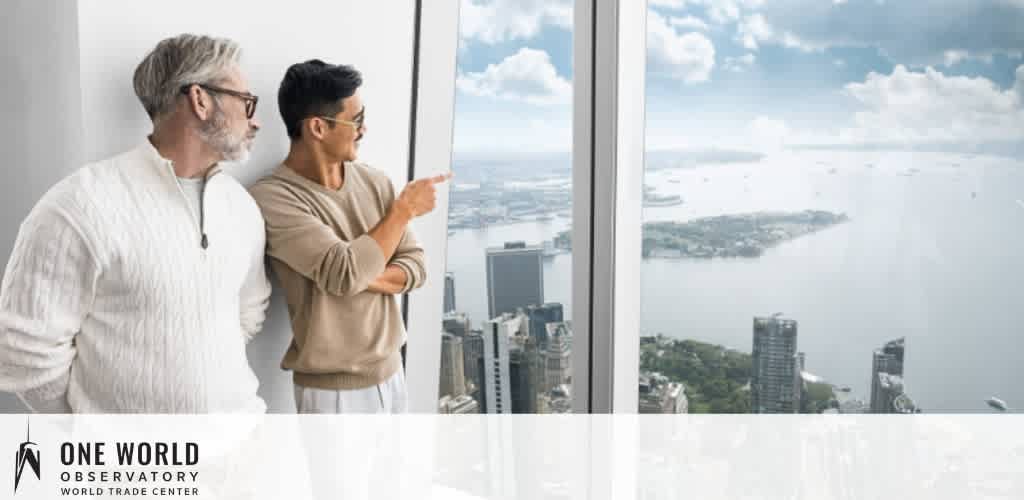 Image shows two individuals standing at a large window inside the One World Observatory. On the left, an older man with gray hair and glasses joins a younger man on the right, both looking out over a cityscape with blue waters and scattered clouds above. The observatory logo is visible in the bottom left corner.