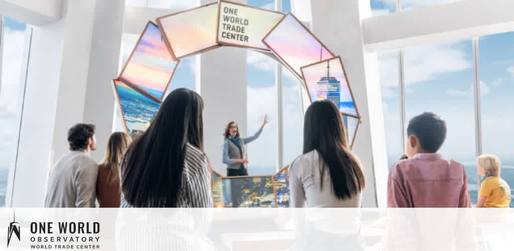 Image showing a diverse group of individuals inside the One World Observatory at the World Trade Center. They are focused on a guide presenting information next to large windows offering an expansive view. The interior has a modern design with bright lighting and digital displays enhancing the presentation.