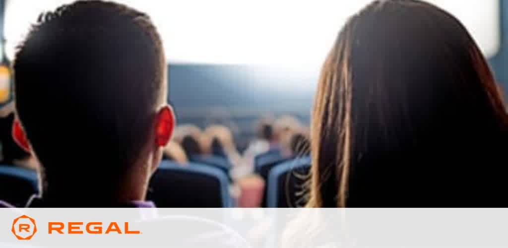 Image shows the back view of two individuals seated at a cinema. They are watching a screen that is not visible, surrounded by other audience members. The person on the left has short hair, while the person on the right has long hair. The Regal Cinemas logo is in the lower right corner, indicating the venue. The setting suggests a movie theater atmosphere.