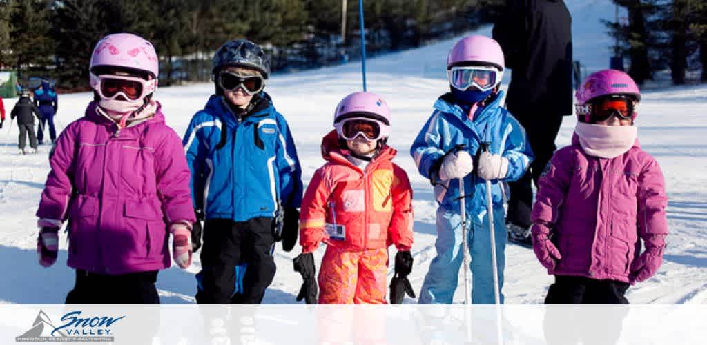 This image shows a group of five young children at a snowy ski resort, presumably Snow Valley, as indicated by the logo in the photograph. They are dressed warmly in colorful ski gear — a combination of pink, purple, orange, and blue jackets. The children are wearing ski helmets and goggles for safety. They appear to be in a skiing lesson or ski school group as they're walking closely together with an adult partially visible in the background. The bright sunlight and clear blue sky suggest favorable weather conditions for winter sports.

At FunEx.com, we're committed to ensuring our customers enjoy their activities with the benefit of the lowest prices available, so everyone can hit the slopes for less!