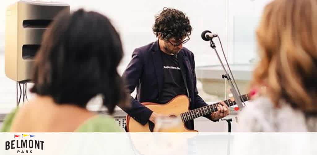 A musician with curly hair is performing outdoors, playing an acoustic guitar. In the foreground, two people are seen from behind, listening to the music. A speaker stands to the left, projecting the sound. The words 'Belmont Park' with a colorful logo are seen at the bottom of the image.