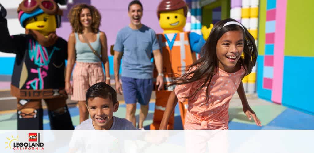 Image of a family enjoying their day at LEGOLAND California. Two children in the foreground are laughing and running towards the camera. Behind them, a man and a woman are walking and smiling, accompanied by two life-sized LEGO characters, one of whom is gesturing a high-five. The backdrop features brightly colored walls representing oversized LEGO bricks.