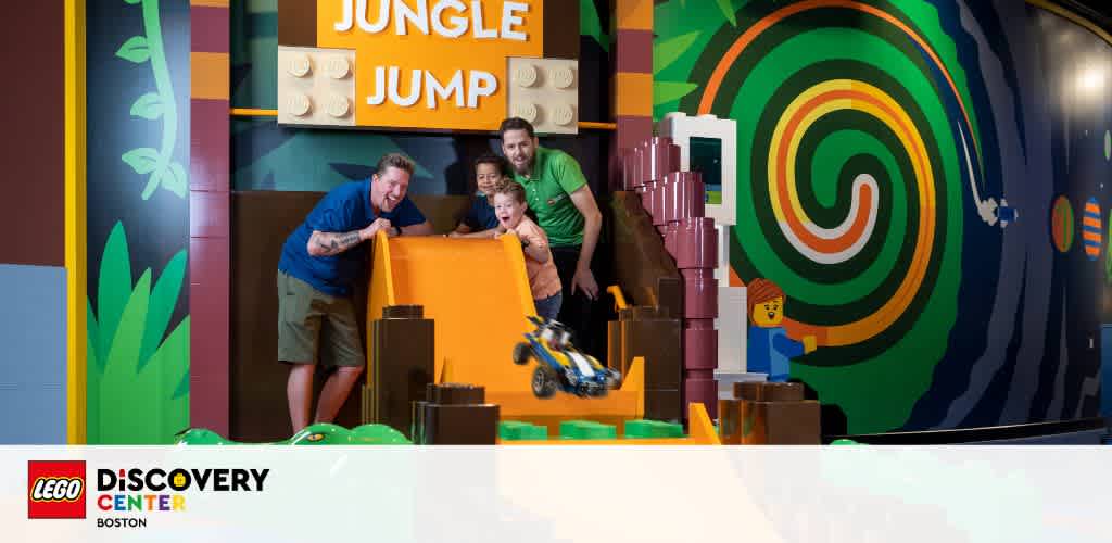 At the LEGO Discovery Center in Boston, three people enjoy a vibrant LEGOLAND Jungle Jump attraction. A thrill of joy is evident as they launch a LEGO car off a ramp amidst large LEGO blocks and colorful jungle-themed decorations.