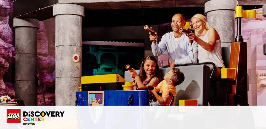 A joyful family of four is engaging in a playful activity at the LEGO Discovery Center Boston. Two children, a girl and a boy, along with their parents, are on a ride with toy guns in hand. They are all smiling and looking towards the same direction, appearing to be in the middle of a lighthearted game. The background features LEGO-themed decorations. The ambiance is vibrant and fun-filled.