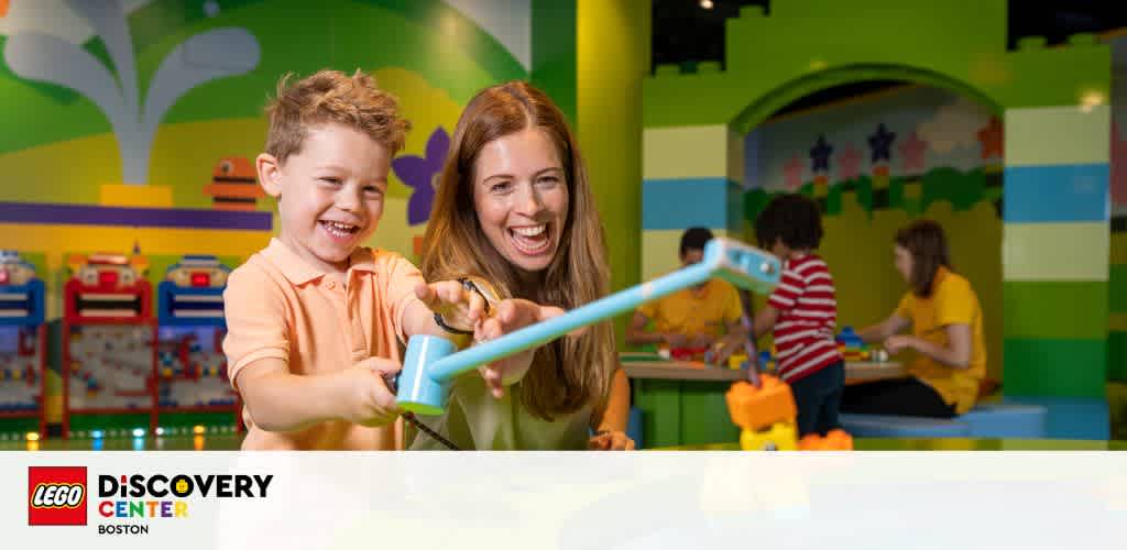 A smiling child and adult play with a toy at the LEGO Discovery Center Boston. Brightly colored blocks and playful decor are in the background. The setting is vibrant, fostering a fun and creative environment for family activities.