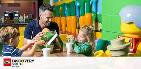 A joyful scene at LEGO Discovery Center Boston where a man is enjoying a meal with two children. They're at a colorful table with oversized LEGO figurine decorations in the background, enhancing the playful atmosphere.