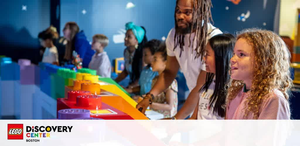 Visitors engage with colorful exhibits at LEGO Discovery Center Boston. Adults and children are seen smiling and focusing on the interactive elements in a brightly lit indoor space. Large LEGO blocks and graphic wall decorations add to the lively atmosphere.