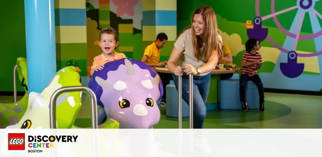 Image depicts a cheerful child and an adult enjoying playful moments at the LEGO Discovery Center in Boston. The adult assists the child on a whimsical amusement ride with colorful LEGO-themed decor in the background, enhancing the vibrant and fun atmosphere.