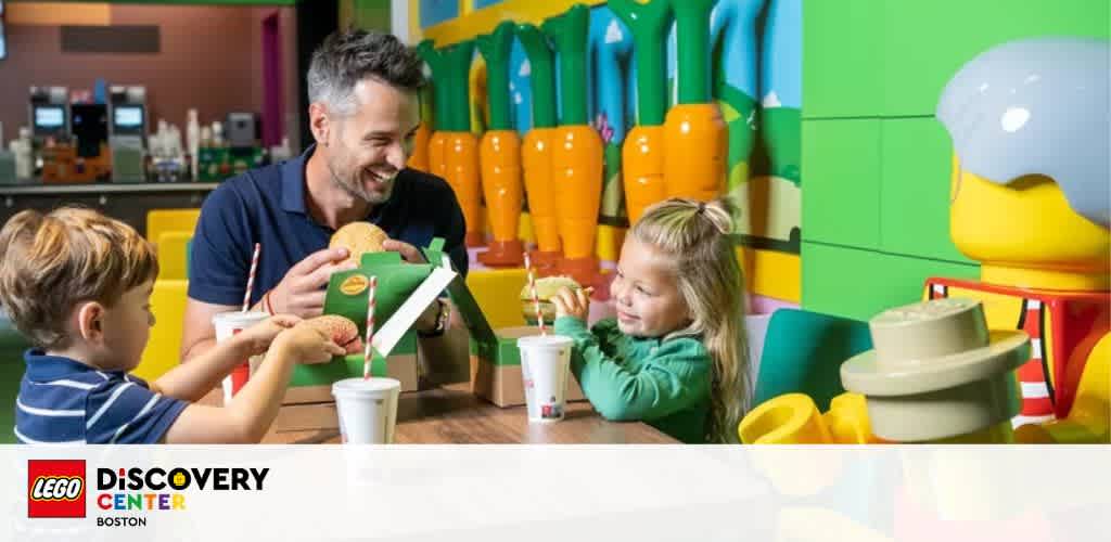 Image shows a joyful moment at LEGOLAND Discovery Center Boston. A man and two children sit at a colorful table, sharing a meal and smiles. Large LEGO figures form the vibrant backdrop.