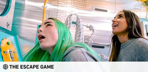 This image features two individuals participating in an escape room activity. The person on the left has vibrant green hair and is looking upwards with an expression of focused curiosity, their mouth slightly open as if in mid-conversation or exclamation. The person on the right has long brown hair and is also looking upwards with a similar expression of engagement and anticipation. Both are surrounded by what appears to be themed decor simulating the environment of the escape game, with noticeable props and gadgets that form part of the challenge. The ambiance suggests a fun and immersive puzzle-solving experience. The logo "THE ESCAPE GAME" is displayed in the lower corner, indicating the brand of the escape room.

At GreatWorkPerks.com, we offer the excitement of such experiences at the lowest prices, ensuring you get the best savings on tickets for your next thrilling escape game adventure.