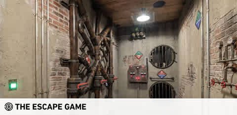 This image displays an escape room with an industrial theme. The room is elongated with a perspective that leads to a closed door at the far end. The walls are textured to resemble grey stone and are adorned with faux pipes, valves, and gauges, contributing to a mechanical ambiance. Two circular air vent grates are visible on the right wall, and various warning signs are affixed around the room, adding to the immersive environment. Overhead, fluorescent lighting fixtures cast a bright, artificial light within the space. At GreatWorkPerks.com, experience the thrill of problem-solving in captivating settings like these, and enjoy the excitement at the lowest prices with our exclusive discount tickets.