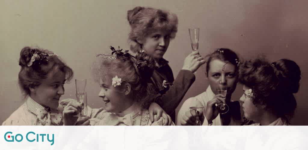 Image displays a vintage photo of five women donning period attire with decorative hairstyles, each engaging in different activities such as holding a champagne flute, peering through a handheld telescope, and conversing with one another. The Go City logo is visible in the bottom left corner. The photo has a sepia-tone color scheme, adding to its historical feel.