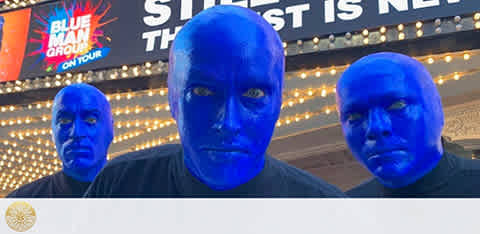 Three individuals with blue-painted faces and bald caps stand in front of a lit sign that reads 'Blue Man Group'. They wear black clothing and have neutral expressions. The sign indicates that the show is now on tour. The background suggests an evening setting with lights.