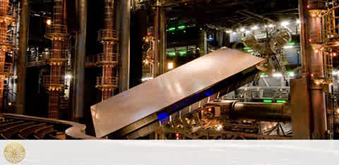 Image depicts an industrial setting with large metal pipes and cylinders, illuminated by ambient lighting. The focal point is a conveyor belt transporting a sheet of material at an inclined angle. The environment suggests a manufacturing or processing plant.