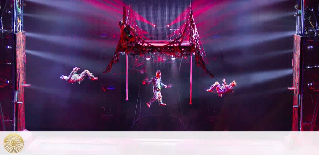 A vibrant circus performance in progress with aerial acrobats performing daring feats. Four performers hang gracefully from a red harness system against a background of dramatic red and purple lighting effects, casting an aura of excitement and energetic entertainment.