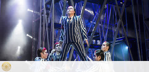 Performers in striped costumes captivate on stage amidst a striking set with metal structures under blue lighting. The central figure stands elevated, commanding attention, while surrounded by attentive co-performers.