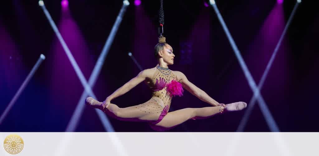 An aerial performer is suspended mid-air in a split position, held aloft by a harness. She's wearing a pink and beige embellished costume. The stage background is illuminated by purple and white lights crossing each other, creating a dramatic environment.