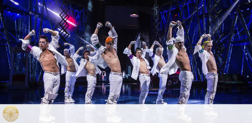 On stage, a group of male dancers with bare chests performs a synchronised routine. They wear white pants, unbuttoned white shirts, and various colored headbands. Some flex their muscles while others hold poses. The backdrop features blue stage lighting and structural stage elements. The atmosphere conveys energy and entertainment.