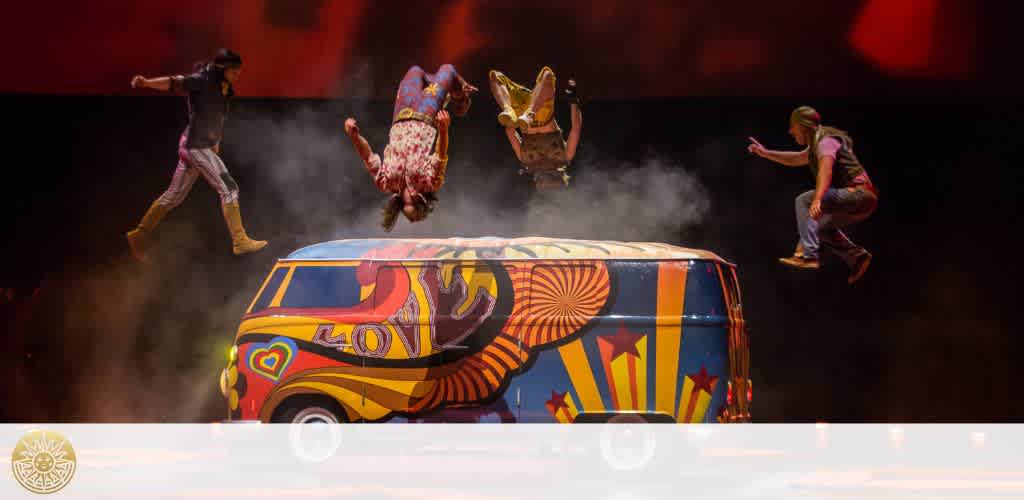 Four acrobats in vibrant costumes leap and flip over a colorful, patterned van on stage amidst a wash of red lighting and hazy effects, giving a sense of dynamic motion and a lively, circus-like atmosphere.