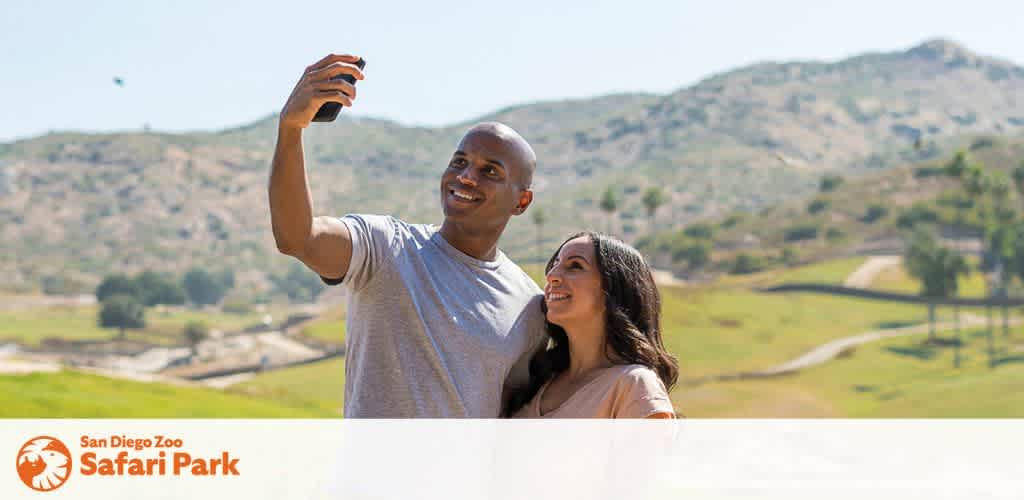 This image features a smiling man and woman taking a selfie together. The man, who is bald and wearing a light gray t-shirt, is holding the smartphone to capture the moment. The woman, with long dark hair, is wearing a peach-colored blouse and is looking up at the man with a joyful expression. They are standing in front of a picturesque landscape that showcases the rolling hills and natural habitats of the San Diego Zoo Safari Park under a clear blue sky. The park's logo is visible in the lower-left corner of the image, signifying the location where the photo was taken.

Experience the thrill of the San Diego Zoo Safari Park and capture your own memorable moments while enjoying savings with our exclusive discount tickets, offering you the lowest prices for your next adventure.