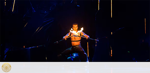 Image shows a performer in dynamic lighting, centered on stage with bare torso, partaking in a fire dance. The dancer has two flaming torches, creating a visually captivating scene against the dark, enigmatic background punctuated by glowing embers.