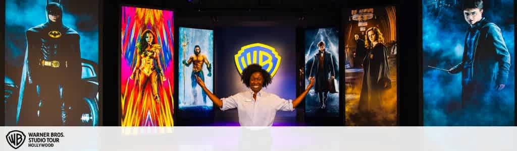 This image displays a joyful person with arms outstretched, standing in front of a series of colorful and vibrant posters from Warner Bros. movies. From left to right, the posters depict iconic characters: Batman, Wonder Woman, Aquaman, and Harry Potter characters Harry, Hermione, and Ron. The Warner Bros. Studio Tour Hollywood logo is prominently displayed in the center, symbolizing the exciting studio tour experience one can anticipate. In one corner of the image, the logo reassures visitors of the authentic and official nature of the tour.

For a magical day out without making your wallet disappear, FunEx.com offers exclusive discounts and savings on tickets to experiences like the Warner Bros. Studio Tour Hollywood, ensuring you always get the lowest prices.