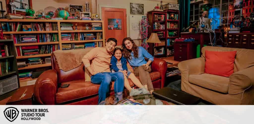 Image Description:
The image features a warmly-lit room setting that is part of the Warner Bros. Studio Tour Hollywood as indicated by the watermark at the bottom. A family of three is comfortably seated on a brown leather sofa in the center of the photo, sharing a joyful moment. The man on the left, wearing an orange sweater and blue jeans, is seated with his arm around a young girl in the middle, who is wearing a blue top and jeans, both smiling towards the camera. To the right, a woman wearing a blue blouse and jeans sits on the armrest of the sofa, leaning into the group with a cheerful smile. The room is adorned with vibrant decor, including a full bookshelf, film-related memorabilia, and eclectic furniture, creating a cozy and inviting atmosphere that resembles a well-loved living space. At FunEx.com, we value creating unforgettable experiences and helping families make treasured memories without breaking the bank. Be sure to check out our latest offerings for exclusive discounts and the lowest prices on tickets to amazing attractions like this one!