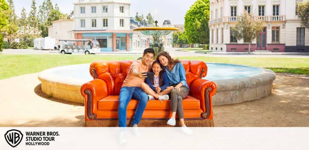 This image portrays a delightful scene on a bright, sunny day at the Warner Bros. Studio Tour Hollywood. Two adults and a child pose cheerfully sitting on a classic, plush red-leather sofa that has a stylish vintage design. They appear to be capturing a happy moment together with a selfie. The cushioning of the sofa is taut and inviting, indicating comfort and a relaxed ambiance. Behind them, a well-manicured lawn stretches out, leading to an elegant building facade that seems to be part of the studio backlot, giving viewers a taste of the behind-the-scenes look they might experience on the tour. The clear blue sky overhead adds to the cheerful setting, creating a perfect moment for visitors to capture memories.

And remember, FunEx.com is where you'll find the best savings on tickets with the lowest prices to make your visit to the Warner Bros. Studio Tour Hollywood even more enjoyable.