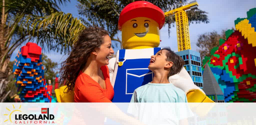 Image shows two happy individuals in an amusement park. A large Lego figure stands in the background between colorful brick sculptures and rides, with the Legoland California logo at the bottom. Bright skies and greenery surround them, suggesting a sunny, fun-filled day out.