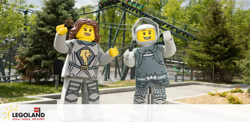 Image shows two oversized LEGO minifigure statues in a park setting. On the left, a figure with long hair and medieval attire, right hand raised. On the right, a knight with a helmet and a smiling expression. In the background, leafy trees and a roller coaster structure are visible, with the  LEGOLAND New York Resort  logo at the bottom.