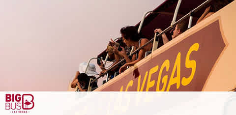 Image of people on the upper deck of a Big Bus Las Vegas tour bus. They appear engaged and are taking pictures. The city ambiance and clear skies suggest a pleasant sightseeing experience. The bus branding is visible on the side.