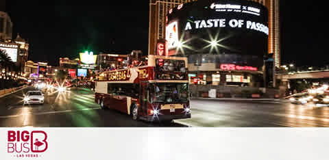 Image shows a Big Bus touring at night along a bustling city street with bright lights and signage. The scene captures a lively urban atmosphere with a sense of motion.