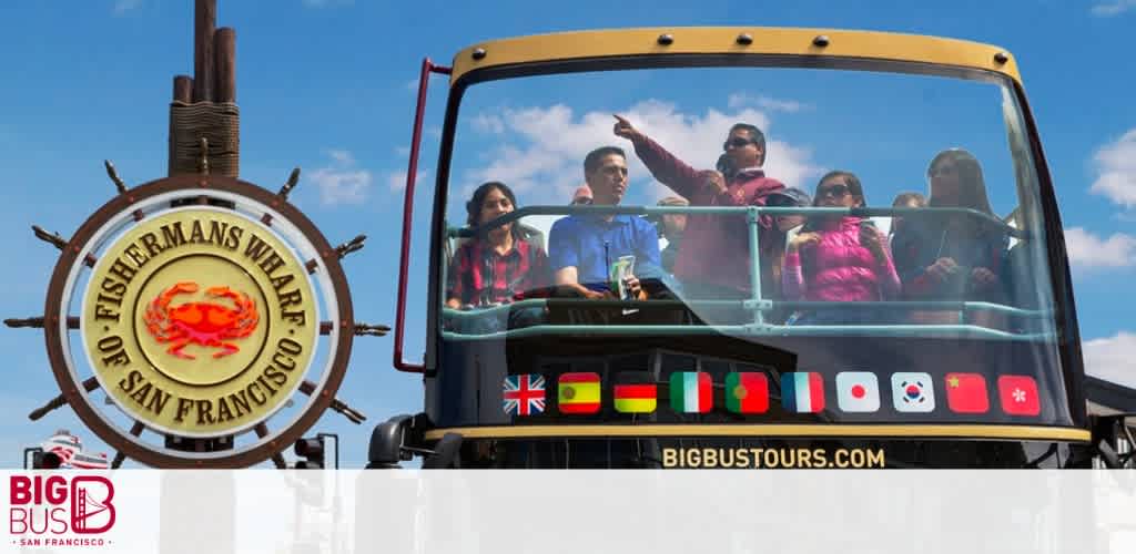 Image shows a group of people enjoying a sightseeing tour on an open-top double-decker bus. To the left, a Fisherman's Wharf sign is prominent. The side of the bus displays the Big Bus Tours logo and website with a row of international flags below. Clear skies suggest good weather for touring.